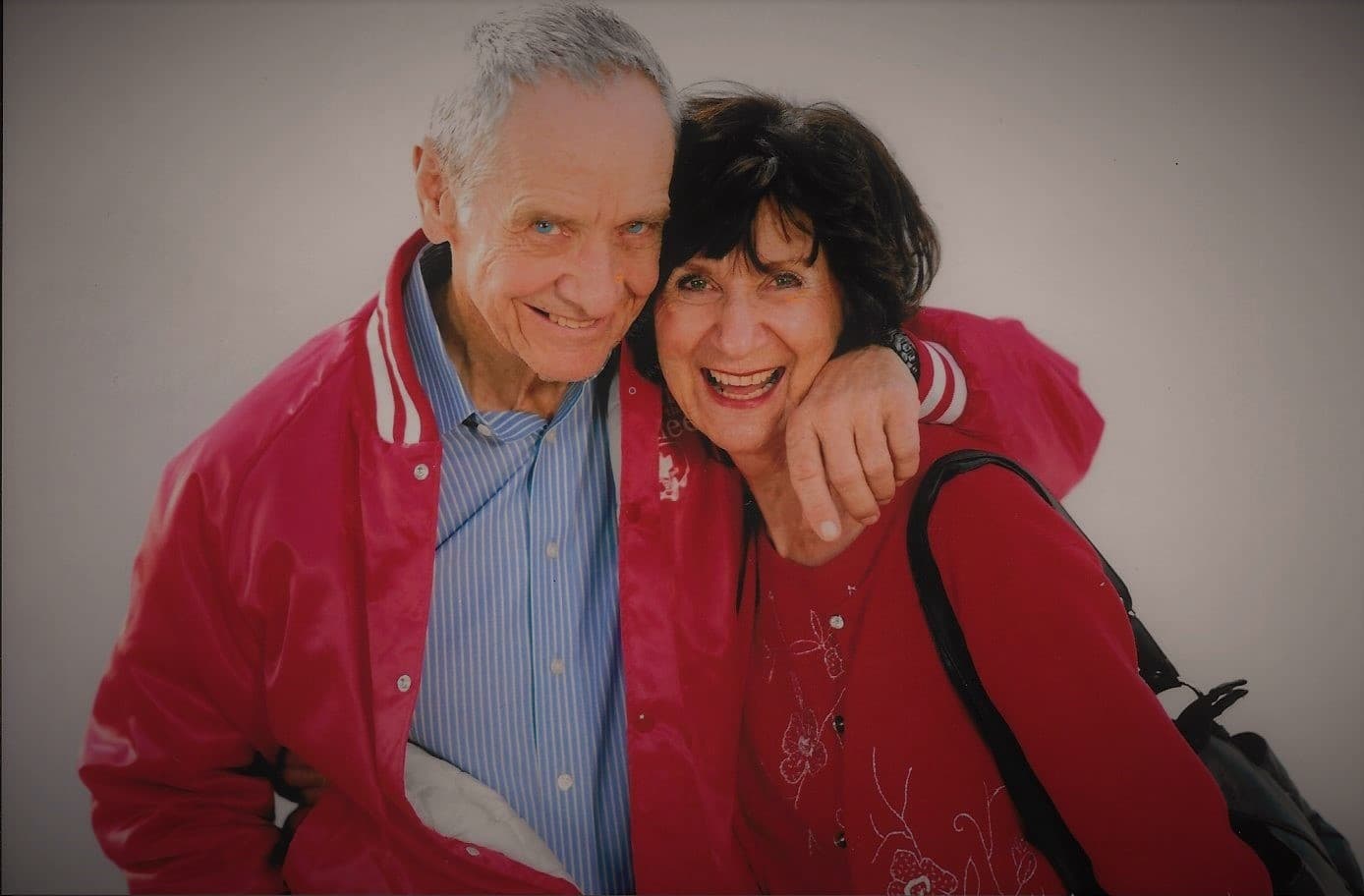 An elderly man and woman smile warmly while hugging, both wearing bright red clothing.