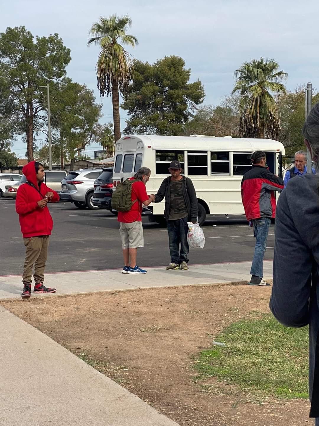 Two men shake hands in a parking lot near a white bus and palm trees.