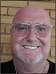 Smiling bald man wearing clear glasses and a grey goatee against a tan brick background.