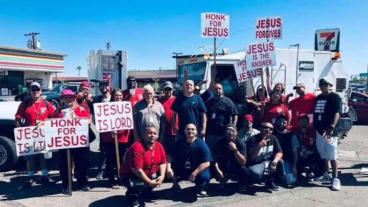 People holding religious signs like 'Honk for Jesus' pose in a sunny parking lot.