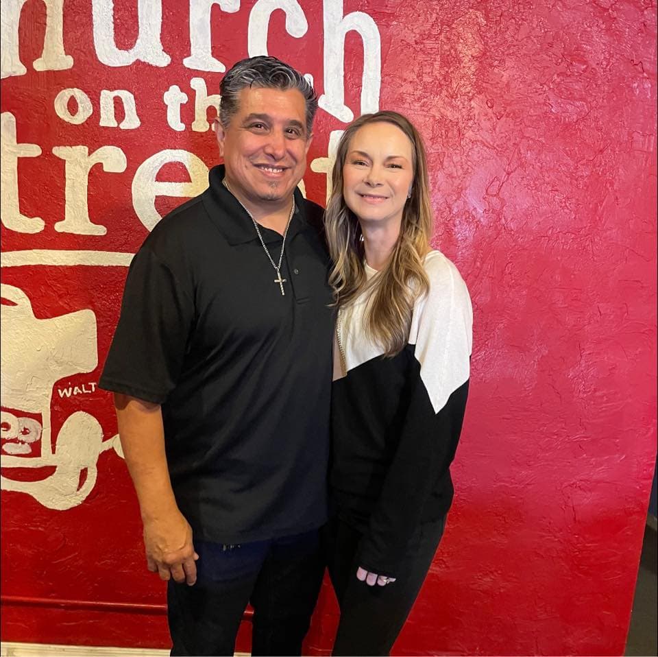 Smiling man and woman pose in front of a red wall with white church lettering.