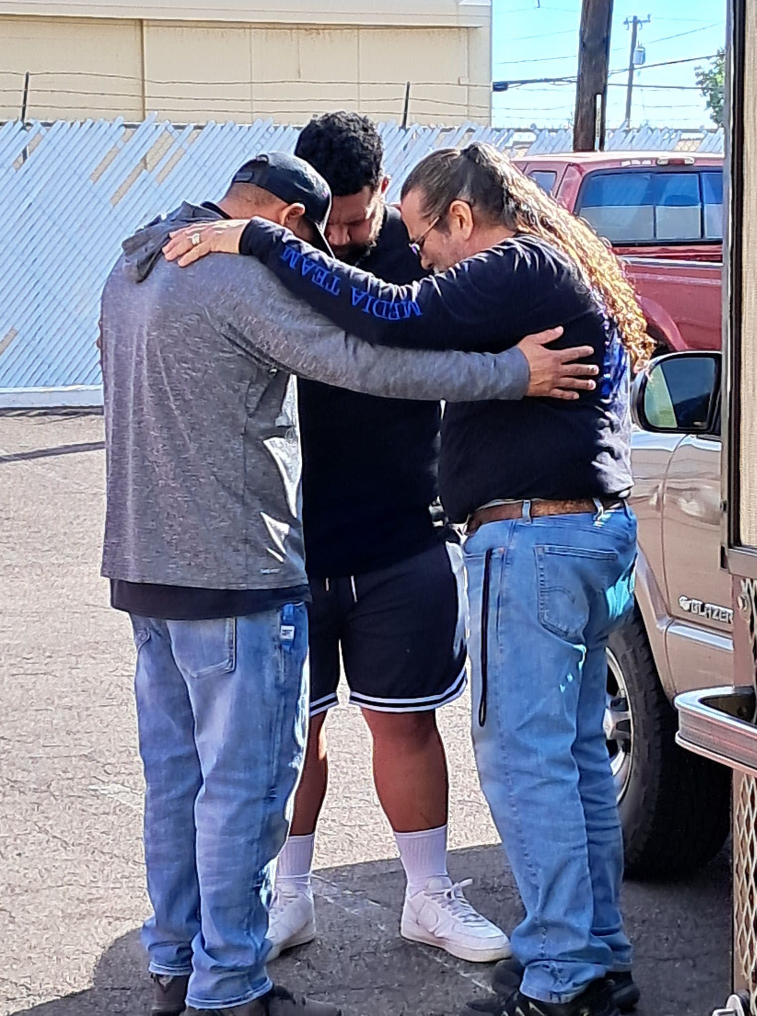 Three men stand in a circle with arms around each other, heads bowed in prayer.