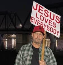 Man in red cap and plaid shirt holding "JESUS LOVES EVERYBODY" sign at night.