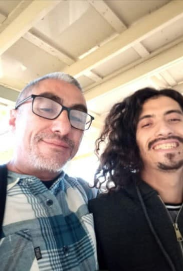 Two men smiling together, one with glasses and another with long, dark curly hair.
