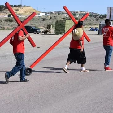 Men in red shirts carry large red crosses with wheels across a paved outdoor lot.
